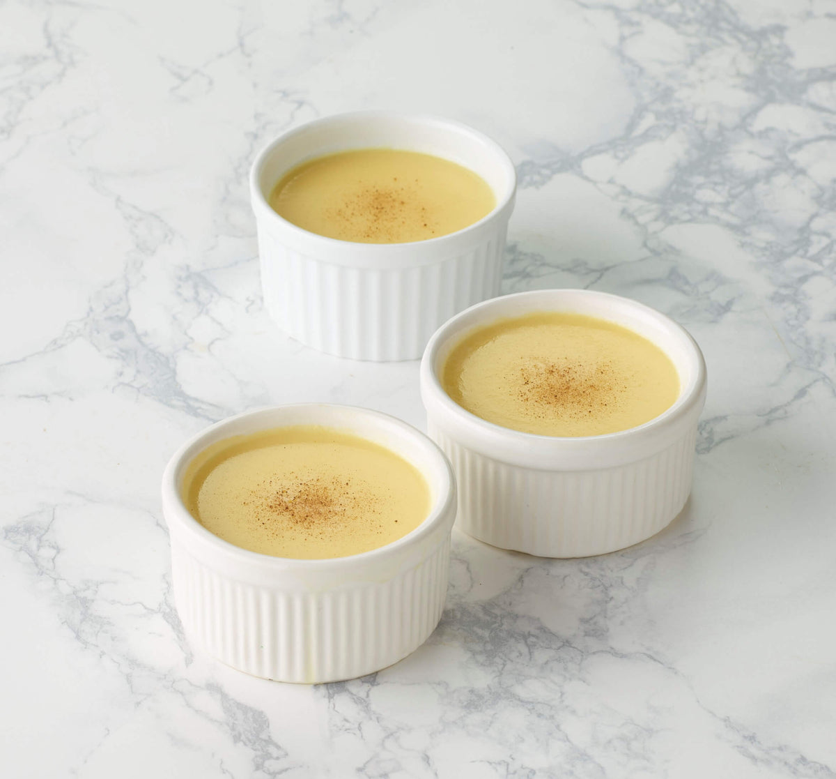 Three white ramekins filled with a yellow custard dessert on a marble surface.