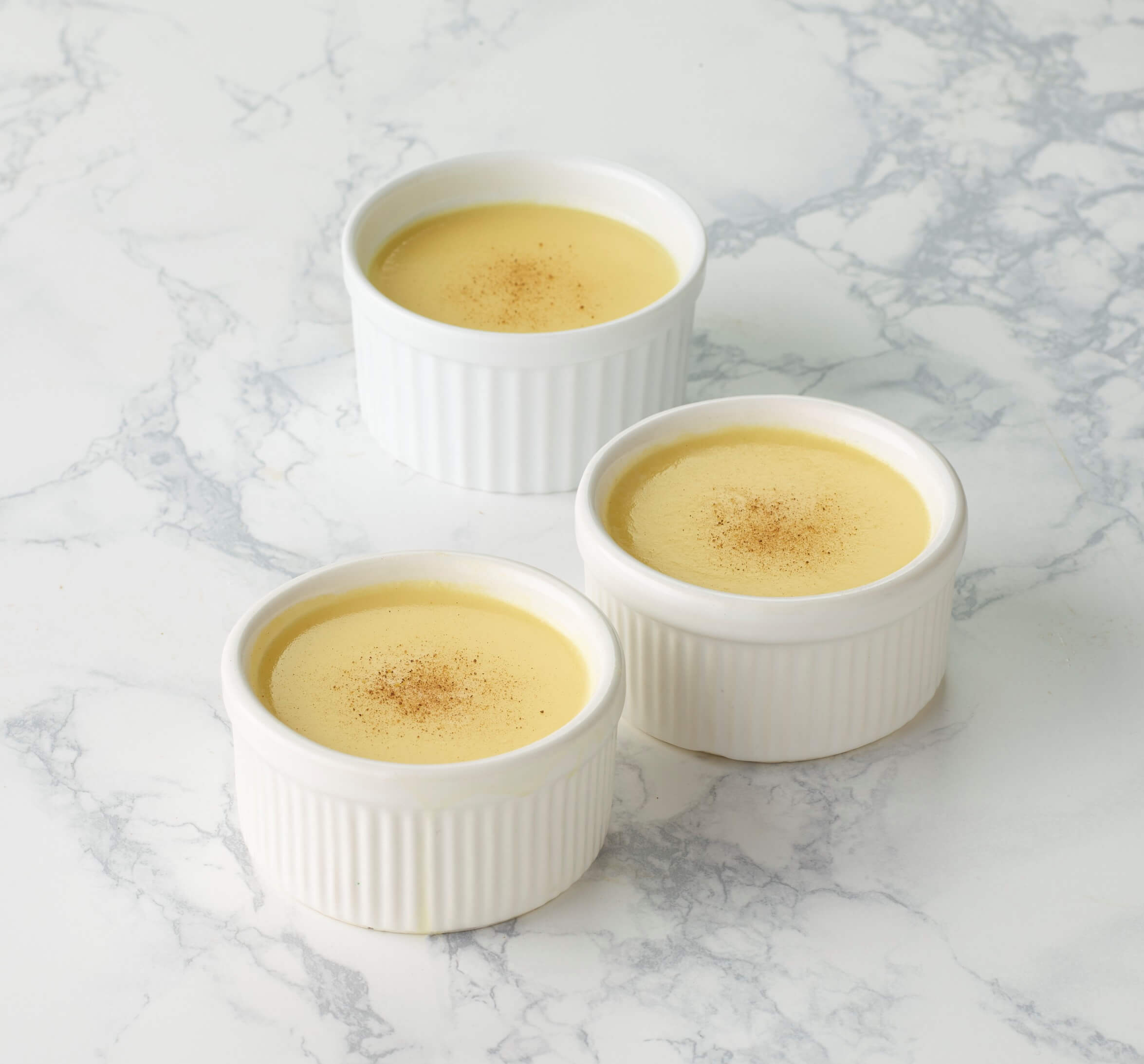 Three white ramekins filled with a yellow custard dessert on a marble surface.