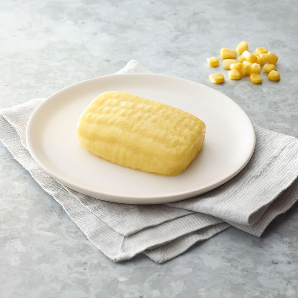 Block of corn on a white plate with corn kernels on a gray surface.