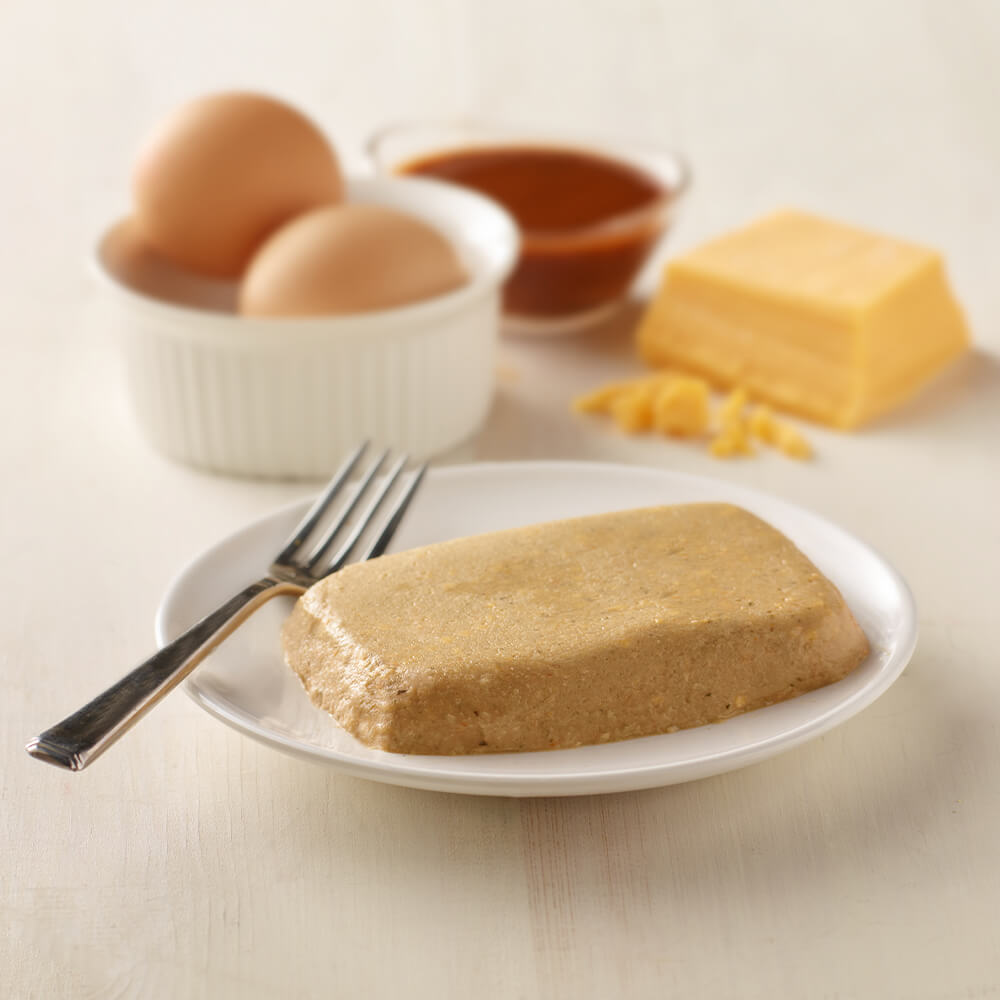Block of brown food on a white plate with eggs, butter, and ketchup in the background.