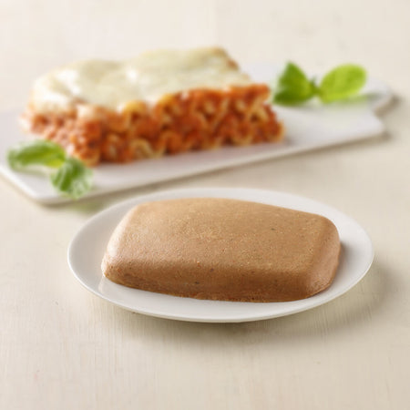Brown rectangular cake on a white plate with a blurred dish of pasta in the background.