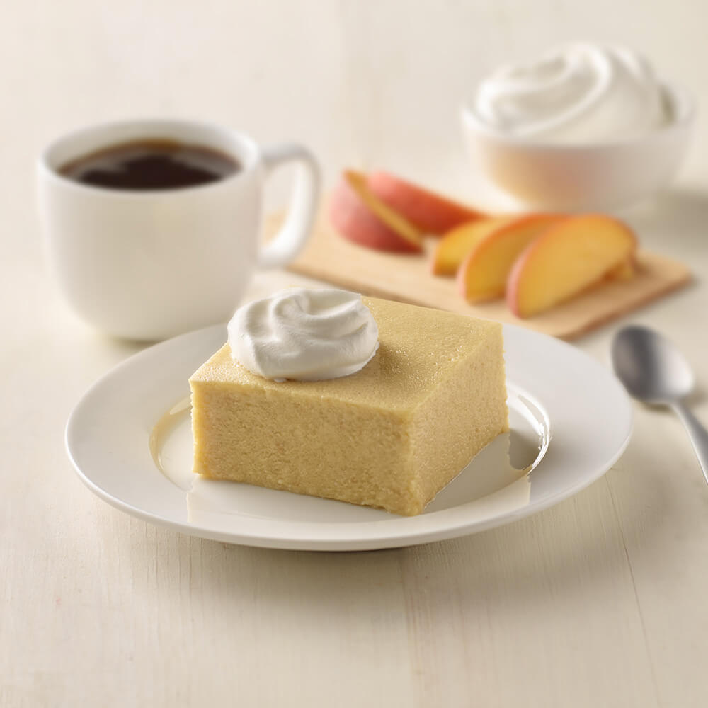 Square piece of peach cake with whipped cream on a plate with a cup of coffee and peach slices in the background.