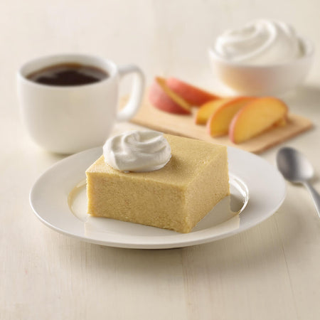 Square piece of peach cake with whipped cream on a plate with a cup of coffee and peach slices in the background.