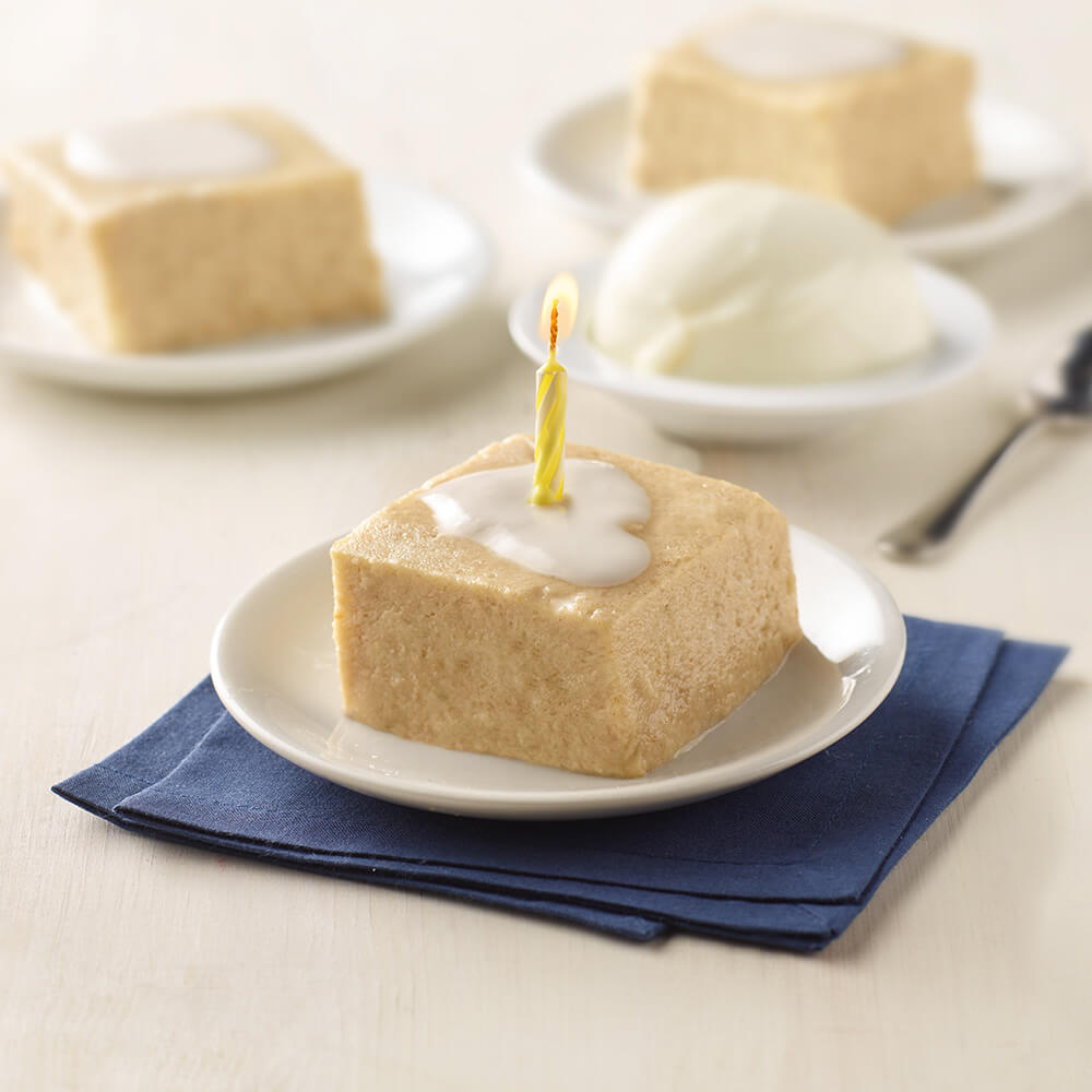 Square piece of vanilla cake with a candle on a white plate with a blue napkin, surrounded by other similar cakes.
