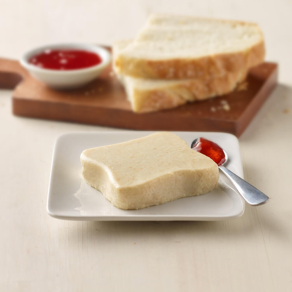 Butter on a piece of bread with a side of jam on a white plate, with more bread and jam in the background.