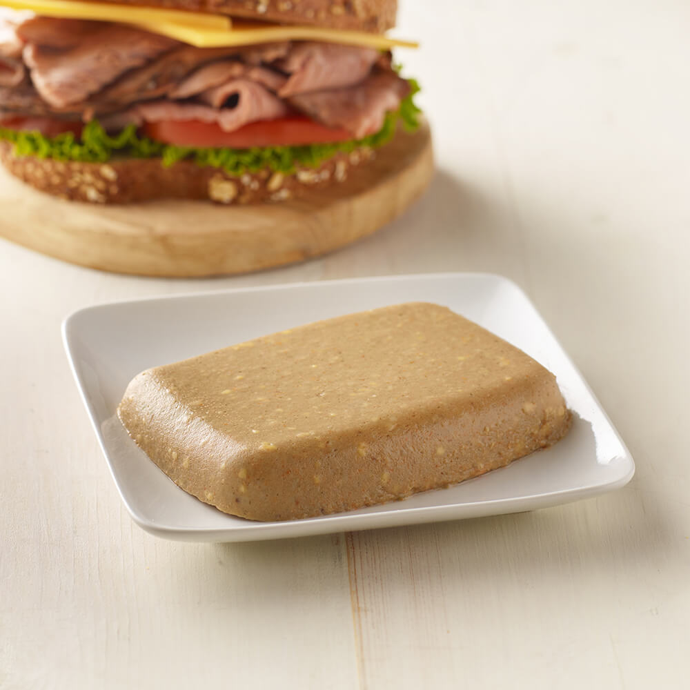Block of brown meat substitute on a white plate with a sandwich in the background.