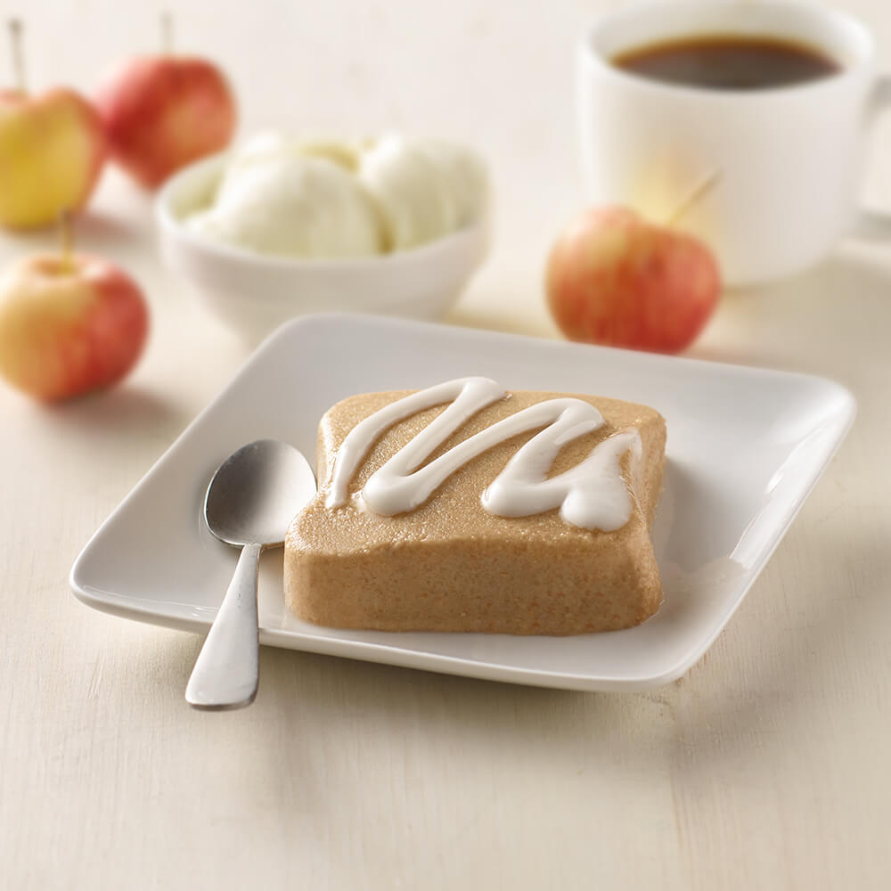 Square piece of apple cake with white icing on a white plate with a spoon, apples, and a bowl in the background.