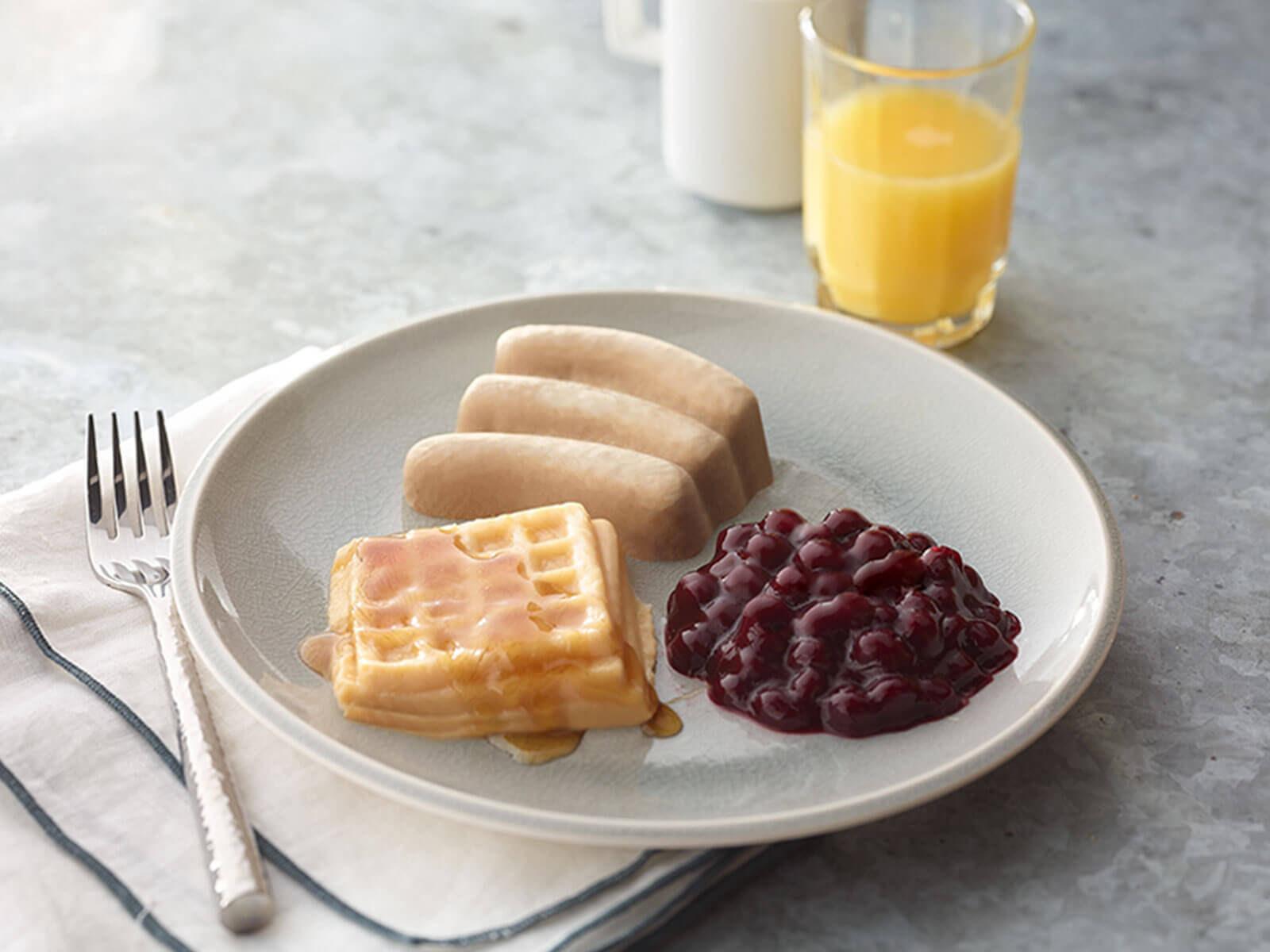 Plate of waffles with syrup, sausage links, and berry compote on a gray surface.
