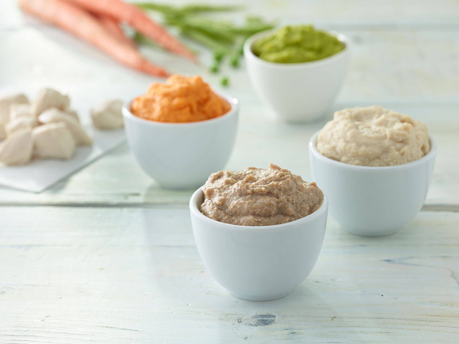 Four white bowls with different types of food on a light wooden surface.